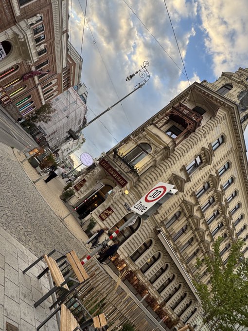 A tilted-angle street photograph of an ornate European-style stone building with multiple stories of arched windows, featuring restaurant signs and a 20 km/h speed limit sign under a partly cloudy sky.