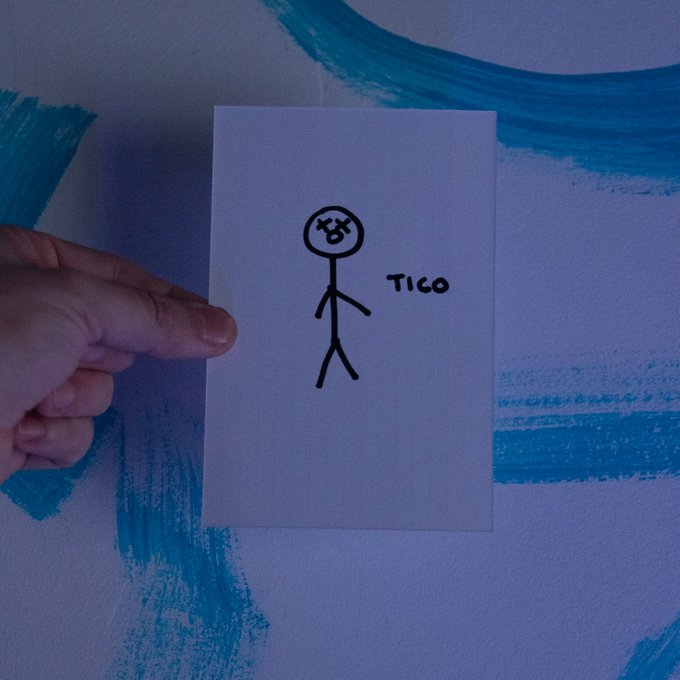 A hand holds a card displaying a hand-drawn angry stick figure with an X for one eye, labeled "TICO," against a wall with blue paint strokes.