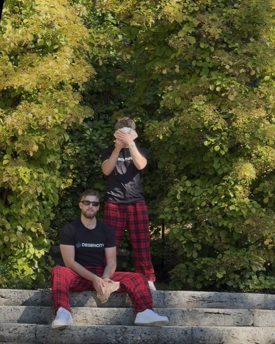 Two young men wearing matching black 'DegenCity' t-shirts and red plaid pants, posing on stone steps with green bushes in the background.