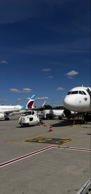 An airport tarmac scene with airplanes parked at gates under a partly cloudy blue sky, featuring a ground service vehicle and orange traffic cone on the runway.