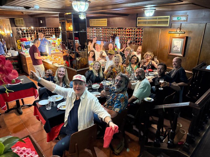 The image shows a group of people gathered around dining tables, enjoying their time together at a restaurant or bar. There are several individuals seated at the tables, with some standing as well. The atmosphere appears lively and social, with everyone engaged in conversation and having a good time. In addition to the people, there is an array of food items on the table, including multiple cups, bowls, and wine glasses. A potted plant can also be seen near one of the tables, adding some greenery to the scene. The overall setting suggests that this gathering could be taking place at a restaurant or bar where patrons are enjoying their meals and drinks in a relaxed environment.