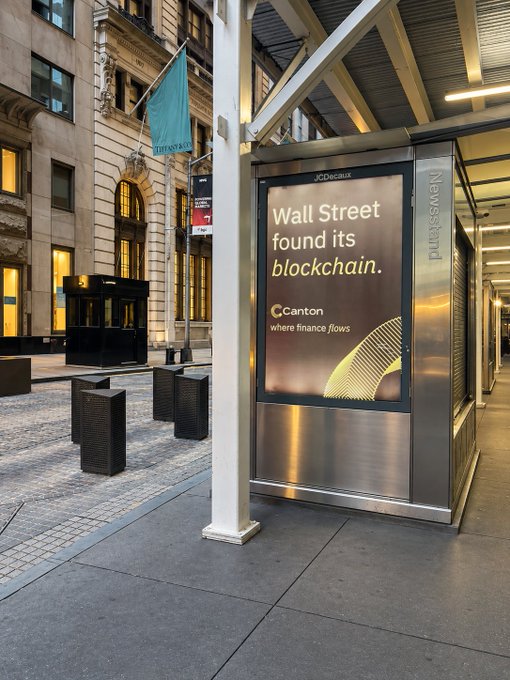 A Canton Network billboard reading "Wall Street found its blockchain. Canton where finance flows" appears at what appears to be a JCDecaux newsstand in a financial district street, alongside a Bernstein flag in the background. The advertisement promotes Canton's blockchain infrastructure for financial markets.