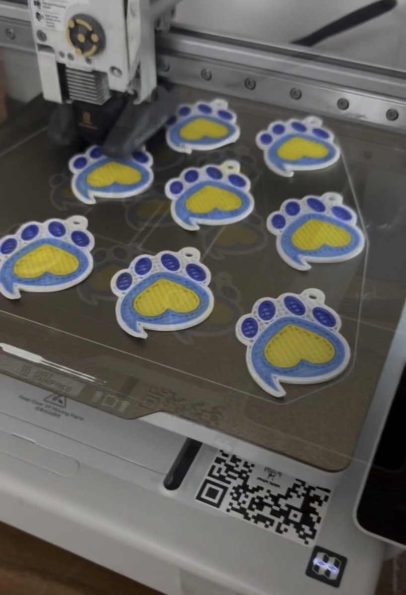 A printer/cutter machine is producing multiple paw-print shaped acrylic charms with blue outlines and yellow centers on its work surface.