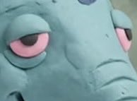 A close-up shot of a green, possibly claymation or animated character's face with large pink-rimmed eyes.