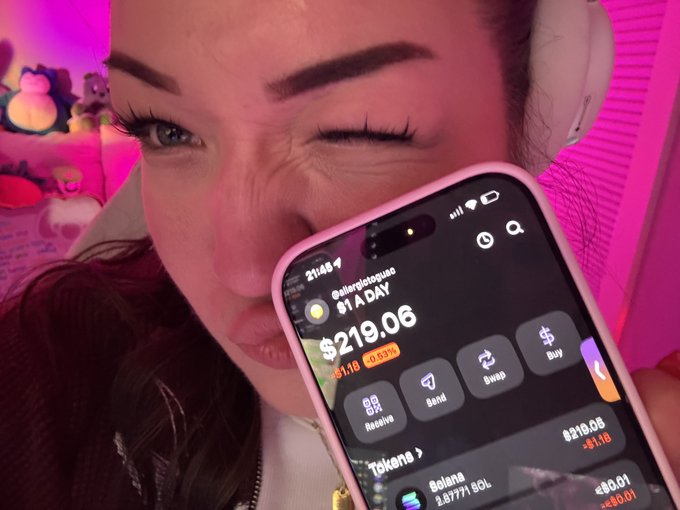 A close-up photo of a person holding a pink smartphone displaying the $AADAY crypto token price of $219.06, showing options like Receive, Send, Swap, and Buy, with Solana (SOL) also visible in the portfolio.