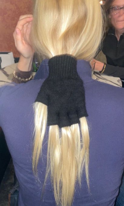 Photo showing the back of a person with long blonde hair wearing a black knitted neck warmer/scarf that has long tassels resembling blonde pigtails hanging down.