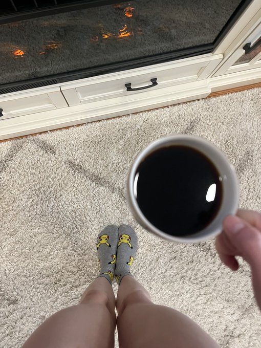 A first-person photo of someone wearing yellow smiley-face socks sitting by a fireplace, holding a white mug of black coffee.