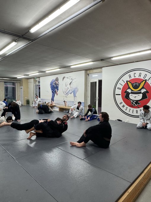 Several people in martial arts gis (mostly black gis) sitting and lying on gray training mats at BJJ Arcade Amsterdam, with two large murals of jiu-jitsu practitioners visible on the back wall.