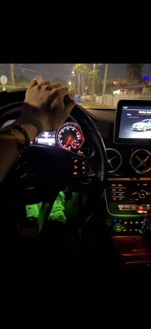 A hand rests on the steering wheel of a Mercedes-Benz at night, with green ambient lighting illuminating the footwell and the dashboard display showing a car graphic. The scene suggests someone preparing for a drop or distribution, aligning with the post about an upcoming address reveal for a $SOL giveaway.