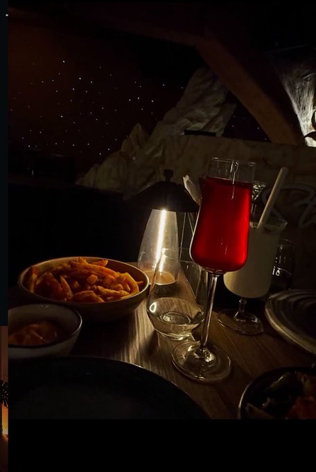 A dimly lit dinner table scene featuring a red cocktail, a creamy drink, a bowl of fries, and a small lamp, with a starry night sky decoration visible in the background.