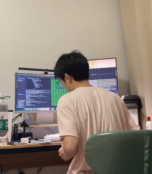 A man with dark hair wearing a pinkish t-shirt sits at a desk working on a computer with dual monitors displaying code, with a water bottle on the desk.