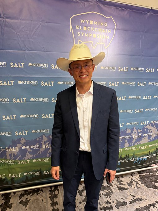A man wearing a cowboy hat, glasses, and a suit smiling in front of a 'Wyoming Blockchain Symposium' backdrop featuring SALT and Kraken logos.