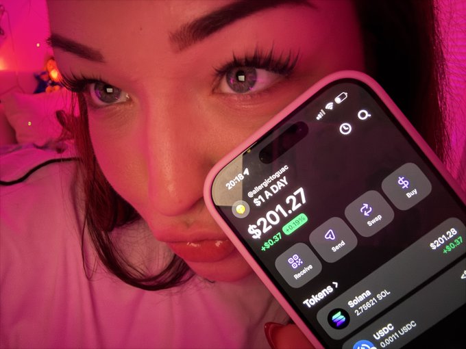 A close-up selfie of a woman under pink lighting holding up her phone, displaying a crypto wallet app with the handle "@energeticgloria" showing a daily profit of "$201.27" for tokens like Solana (SOL) and USDC.