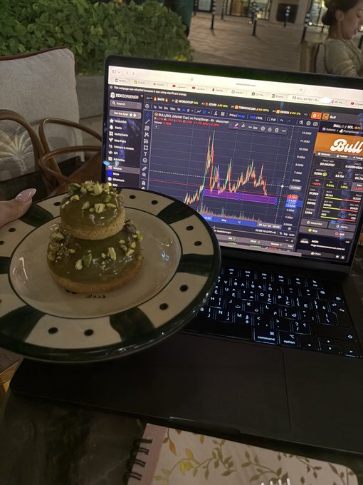 The image features a laptop computer with a plate of food, specifically a green donut with chocolate topping, placed next to it. The background shows a person sitting at a desk or table while using the laptop. The caption of the image reads "Guess who’s gonna touch support soon and fly higher... again? $Bull ish forever". This suggests that someone might be taking a break from work or studying to enjoy their food, possibly discussing the post about the bull market on the computer screen.
