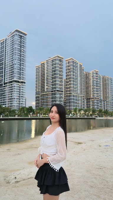 A young woman poses on a sandy beach with high-rise waterfront buildings and a calm canal in the background, fitting the upcoming "Fenie x Ritual #7" collaboration teaser.