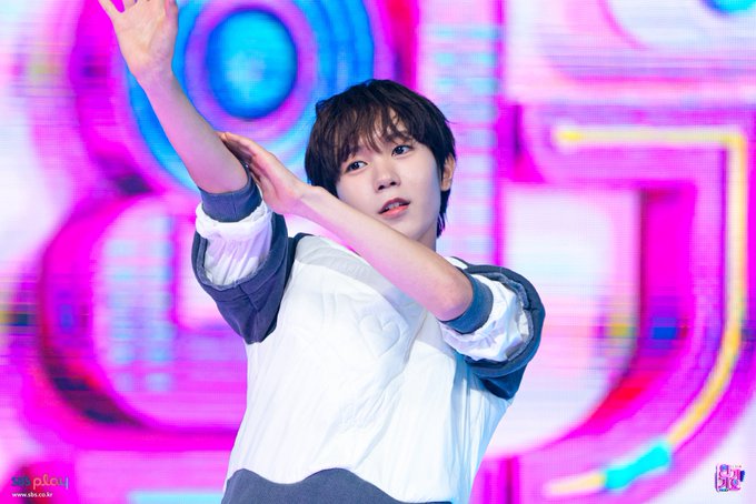 A young male K-pop idol (Jungkook of BTS) striking a dance pose on stage with arms raised, wearing a white shirt over a dark long-sleeve top, set against a colorful, bright pink and blue digital backdrop.