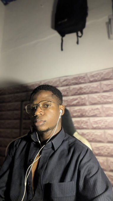 A selfie of a young man wearing glasses, a black striped shirt, and white wired earphones, sitting in a room with a brick-patterned wall.