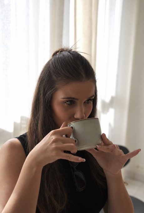 A young woman with long dark hair sips from a gray coffee mug in soft natural light, her gaze directed off to the side. The quiet morning moment with a warm beverage subtly mirrors the post's urgent financial warning—suggesting that simple comforts like this could become luxuries for those who haven't invested in Bitcoin while young.