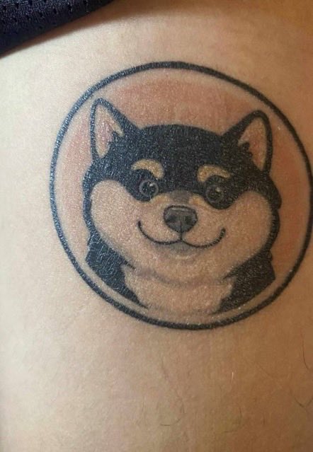 A circular black ink tattoo of a smiling Shiba Inu dog wearing a mask on someone's skin.