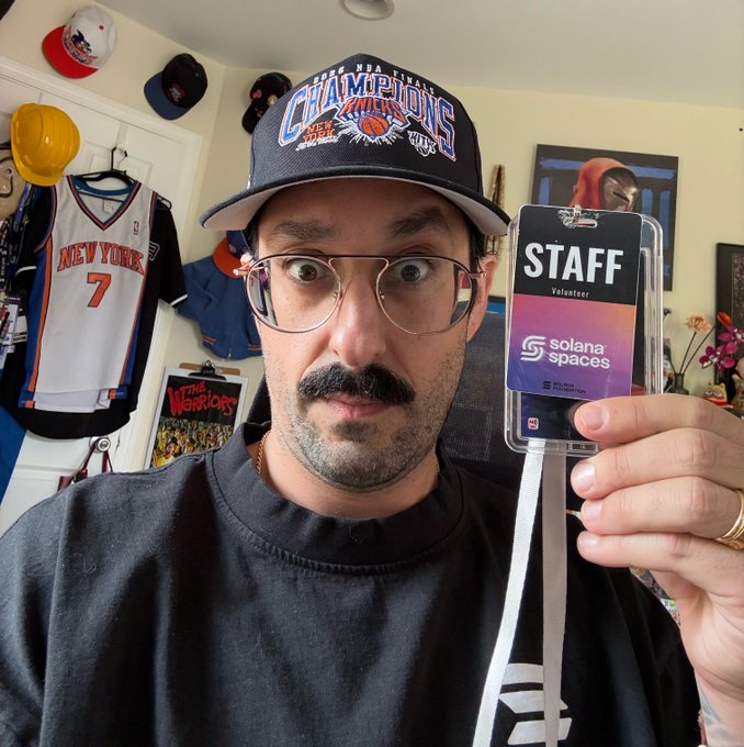 A man with a mustache and glasses, wearing a New York Knicks NBA Finals Champions hat, proudly holds up a "STAFF Volunteer" badge for Solana Spaces while posing in a room decorated with Knicks memorabilia and Warriors gear. The badge connects directly to the post, where he jokes about his role as a dedicated Solana Spaces volunteer for the special event in Los Angeles.