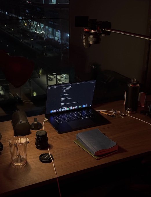 A nighttime desk setup features a laptop displaying code on its screen, accompanied by a camera lens, a drinking glass, notebooks, and a camera mounted on an overhead arm, with a city street visible through the window.