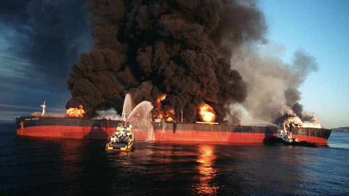 A dramatic scene of a large red oil tanker engulfed in massive flames and thick black smoke on the water, with smaller firefighting boats spraying water onto it.
