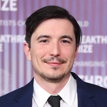 A man with dark hair and a goatee, wearing a navy suit, white shirt, and dark tie, poses in front of a backdrop displaying "BREAKTHROUGH PRIZE" text. The image appears to be from a Breakthrough Prize event and shows no direct connection to the news about Robinhood Chain ($HOOD) surpassing $600 million in deposited assets.