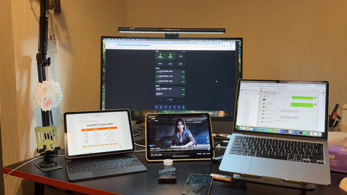 A desk setup displays multiple devices including an iPad, a desktop monitor, a tablet showing a video call, and a MacBook showing a spreadsheet.