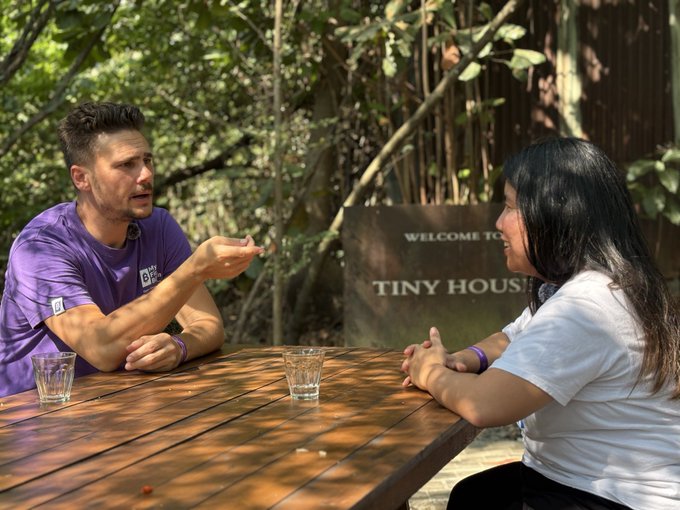 Two people sit at an outdoor wooden table having a conversation, with a sign reading 'WELCOME TO TINY HOUSE' visible in the background.