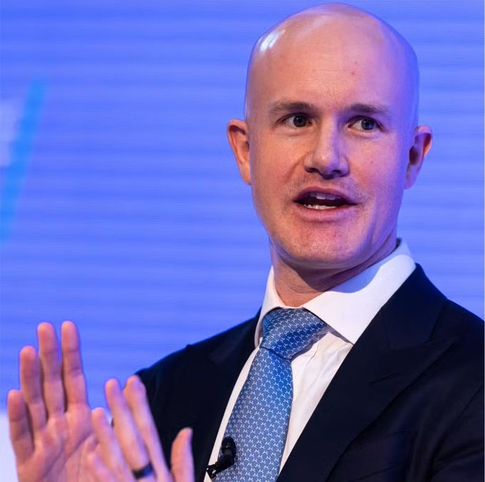 A bald man in a dark suit and blue patterned tie speaking with expressive hand gestures against a blue background.