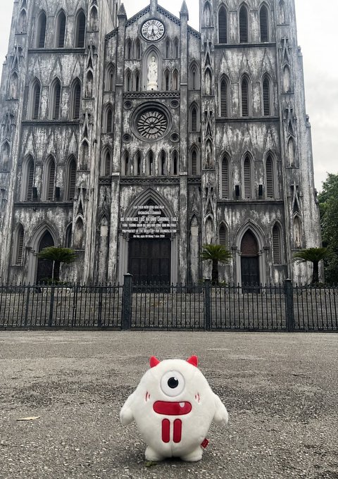 A small white plush toy with red horns and a single eye sits in front of St. Joseph's Cathedral in Hanoi, the striking Gothic Revival facade featuring its clock tower and rose window providing a dramatic backdrop for the travel series mascot.