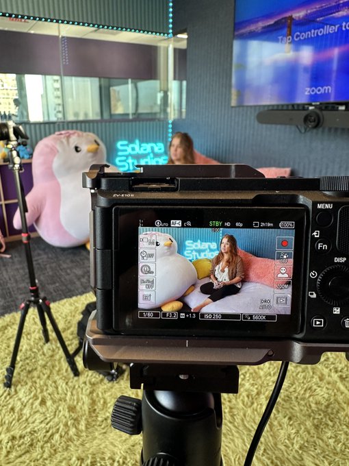 The image shows a camera with a video feed of two people sitting on a couch, possibly recording a cooking tutorial or a live stream. The camera is positioned at an angle that makes it appear as if someone is taking a picture of them from behind. The background features a TV screen displaying the words "Cooking some @RUMSHQ LIVE magic @solana studios".