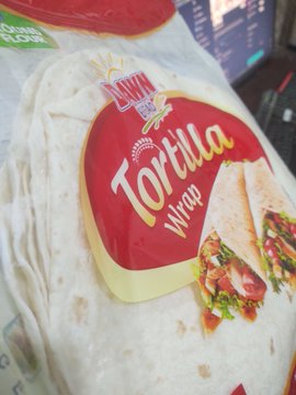 A package of Lazzat Tortilla Wraps sits on a surface, with the brand's red and yellow logo prominently displayed alongside a wrap filled with chicken and salad. The image illustrates the post's message about using a Bitget Wallet Card to pay 598 PKR for everyday grocery items, highlighting how crypto is being integrated into routine purchases.