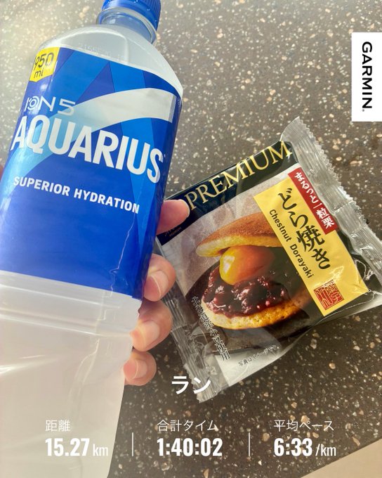 A hand holding a bottle of Aquarius ions Superior Hydration sports drink and a Premium chestnut dorayaki pastry package, with Garmin running stats showing 15.27 km, 1:40:02, and 6:33/km pace.