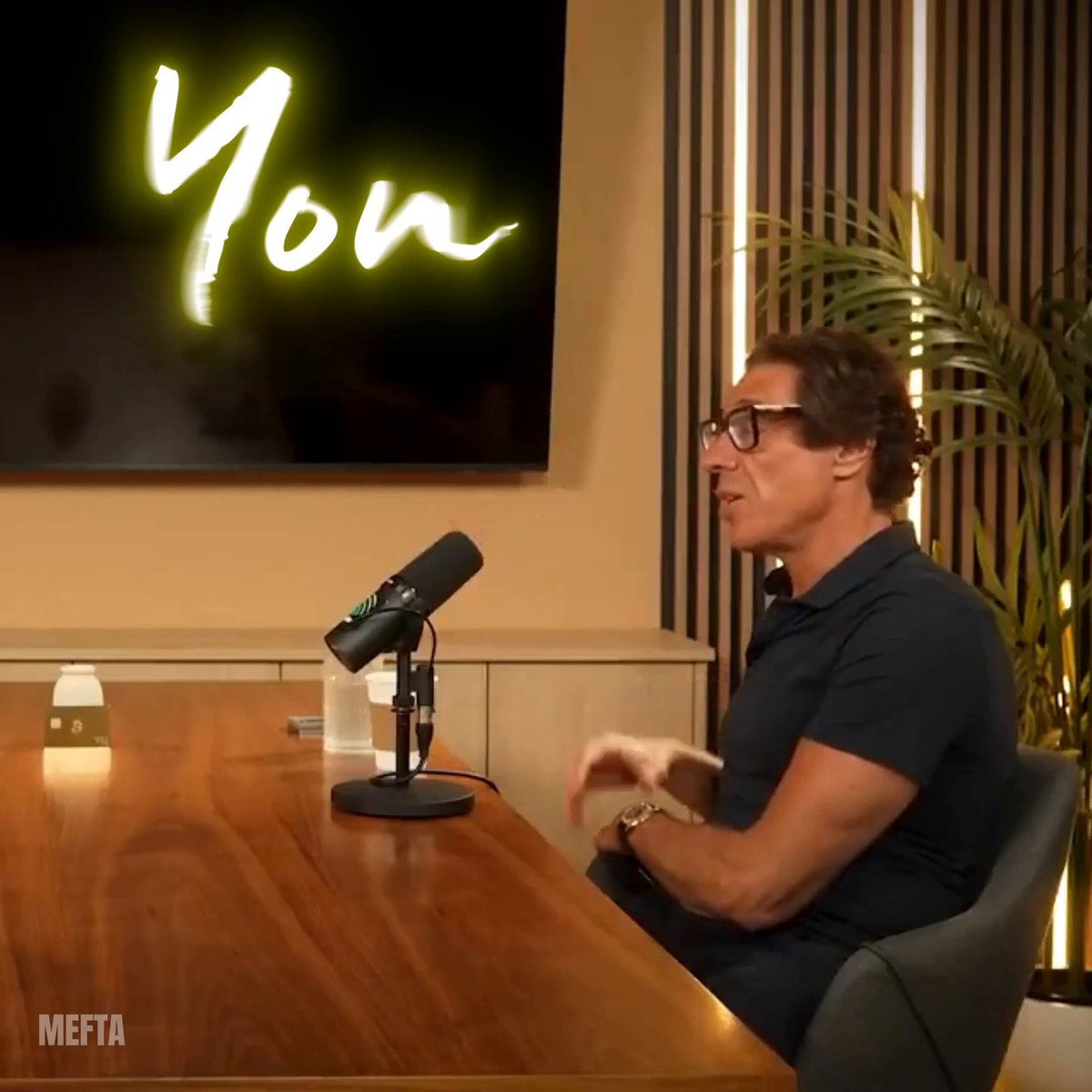 The image features a man sitting at a wooden table, wearing glasses and a black shirt. He appears to be engaged in conversation or giving a speech, as indicated by his body language. The background of the image includes a TV screen displaying the words "YOUR" written on it, along with a plant placed nearby.