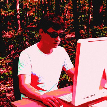 A stylized photo with heavy red and pink color saturation shows a person wearing sunglasses working on an Apple iMac computer against a forest backdrop.