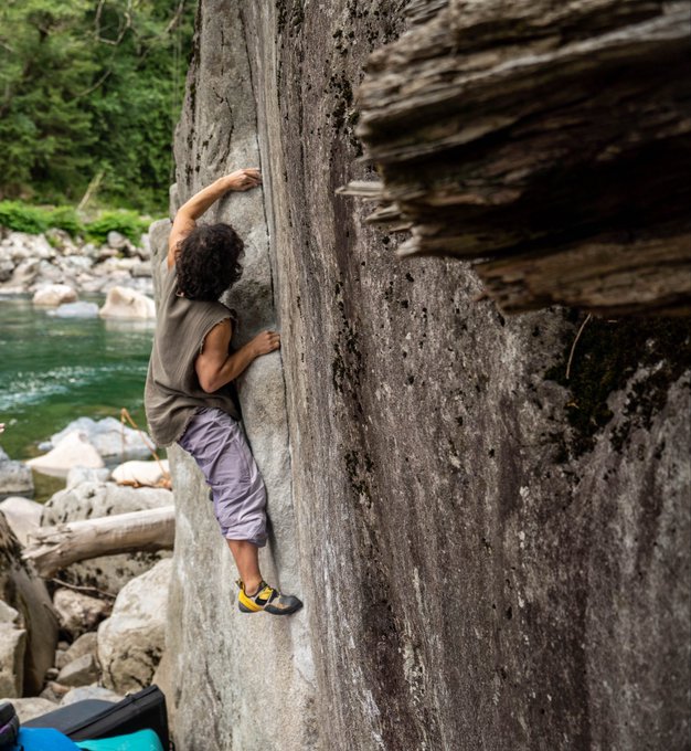 A rock climber scales a towering boulder beside a serene river, gripping the rough stone with focus and determination. The image symbolizes resilience, adventure, and stepping outside one's comfort zone—fitting for the poster's message about staying grounded and engaged with the Azuki community while enjoying life beyond web3.