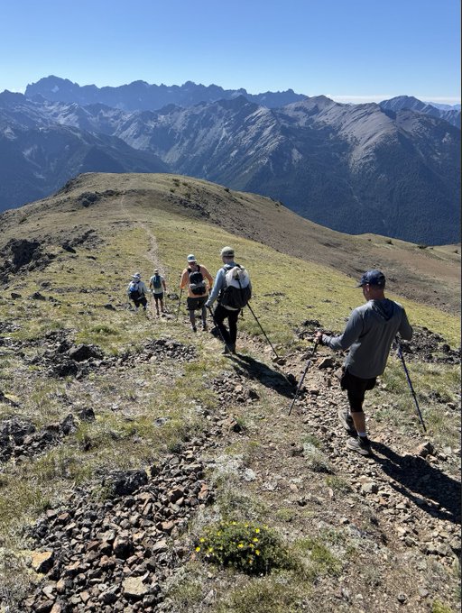 Four hikers with trekking poles walking along a rocky mountain ridge trail, with dramatic snow-capped mountain peaks in the distance.