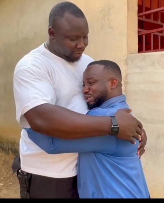 Two men embracing outdoors, with the taller man in a white t-shirt hugging the shorter man in a blue shirt who appears to be crying.