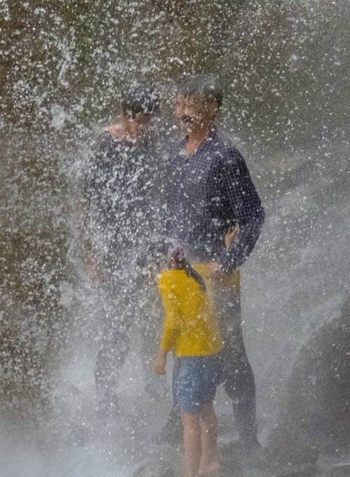 A photograph showing a man and child standing in heavy water spray or splashing water, likely from a fountain or beach wave.