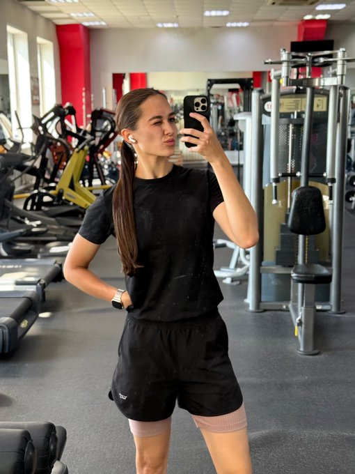 A young woman takes a mirror selfie in a gym wearing a black t-shirt, black shorts, and AirPods, posing with one hand on her hip amid various workout equipment.