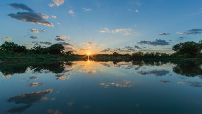 A scenic landscape captures a vibrant sunset reflecting over a calm body of water, framed by silhouetted trees and clouds.