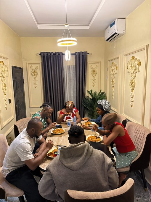 A group of six people gather around a circular dining table sharing a meal together in an elegantly decorated room with ornate wall paneling and a modern pendant light, embodying the post's message about teamwork extending beyond the kitchen into shared mealtime.