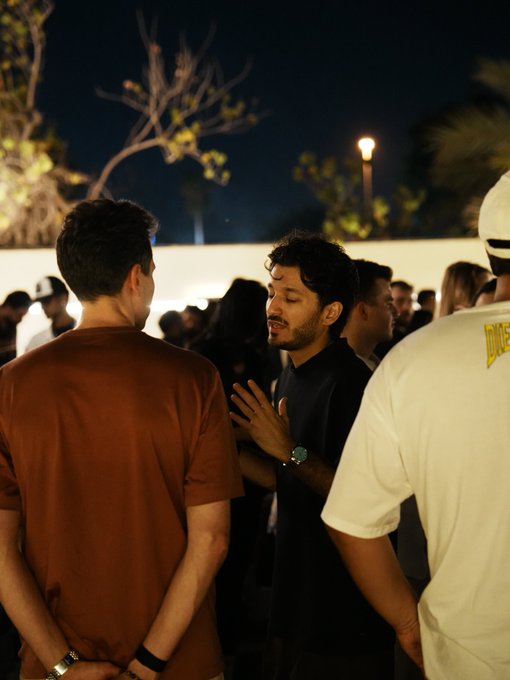 The image shows a group of people standing around each other, with one man wearing an orange shirt and another wearing a white t-shirt. They are engaged in conversation or observing something together. The photo has been edited to include text that reads "me after looking at $BTC price this morning...", which seems to be related to the Bitcoin market.

The image also contains some text about a man's wristwatch, possibly indicating the time of day when the picture was taken.