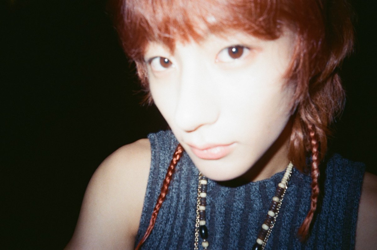 A close-up of a person with reddish hair styled in small braids, wearing a dark sleeveless top and a beaded necklace, looking directly at the camera.