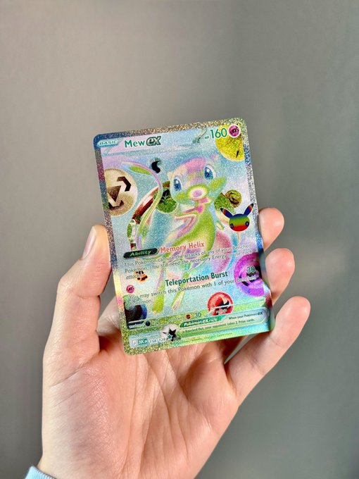A hand holds a holographic "Mew ex" Pokémon trading card with 160 HP, featuring abilities "Memory Hole" and "Teleportation Burst," showcasing the shiny, futuristic design referenced in the caption.