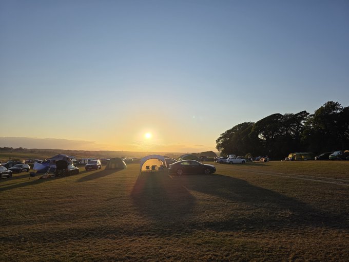 Sunset view over a campsite with tents and cars parked in a grassy field, with trees silhouetted on the horizon.
