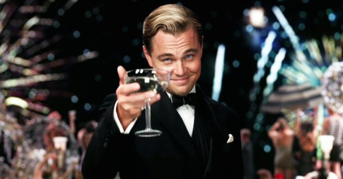 Leonardo DiCaprio, dressed as Jay Gatsby in a tuxedo, raises a champagne glass in a toast amid fireworks and a festive party crowd.
