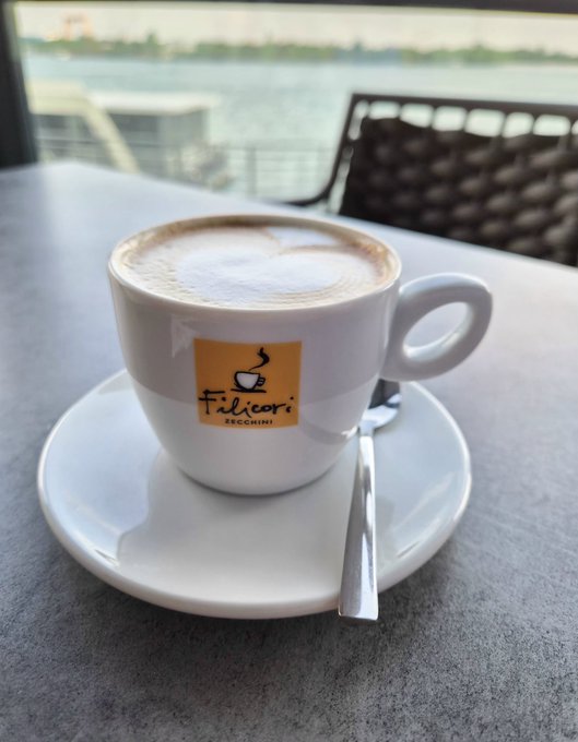 A cappuccino served in a white cup on a saucer at an outdoor table, featuring the 'Filicori Zecchini' logo, with a waterfront view in the background.