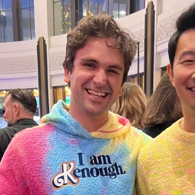 Two smiling men pose closely together, with the central figure wearing a fuzzy, rainbow-colored hoodie featuring the embroidered text "I am Enough." The colorful, optimistic attire and cheerful setting contrast with the biographical caption about Alex Atallah's serious tech career journey from Stanford through Apple and Palantir.